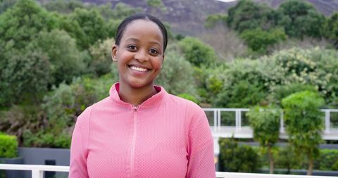 Smiling African American woman standing on balcony wearing pink zip-up jacket amid lush garden