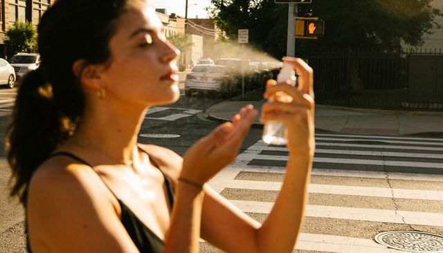 Woman misting face with clear spray bottle at sunlit urban crosswalk