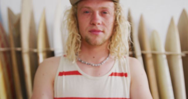 Portrait of caucasian male surfboard maker with long blonde hair in front of surfboards, unaltered. Small business, work, sports equipment and craftsmanship.