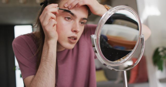 In this image, a young adult is seen intently applying eyebrow makeup before a mirror. Suitable for themes of self-expression, beauty routines, and personal grooming. This stock photo can be used in beauty blog posts, LGBTQ+ publications, self-care articles, and makeup tutorials.