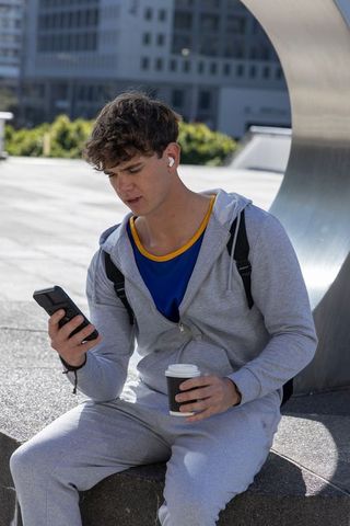 Late teen wearing gray hoodie sitting on ledge checking smartphone while holding coffee