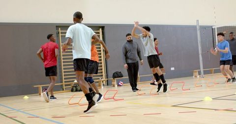 Diverse male athletes doing agility drills in gym with coach observing and guiding
