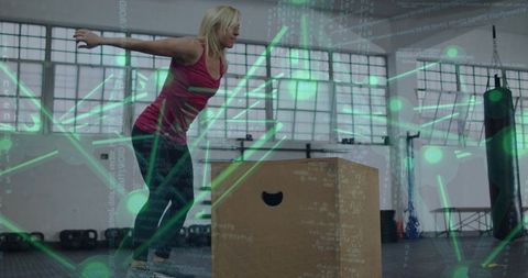 Blonde woman jumping onto plyo box in industrial gym with green digital network overlay