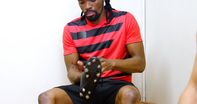 A soccer player in a red and black jersey is sitting in a locker room, putting on his cleats. This image can be used for sports-related advertisements, fitness training guides, promotional materials for athletic gear, or articles focused on pre-game routines and athletic preparations.