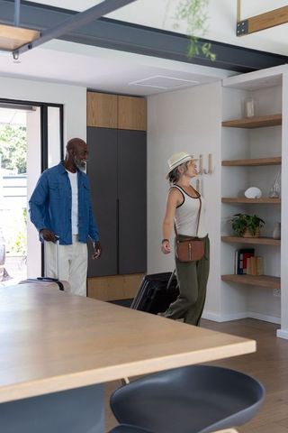 Mixed-race couple arriving at modern home with wheeled luggage, exploring bright entryway