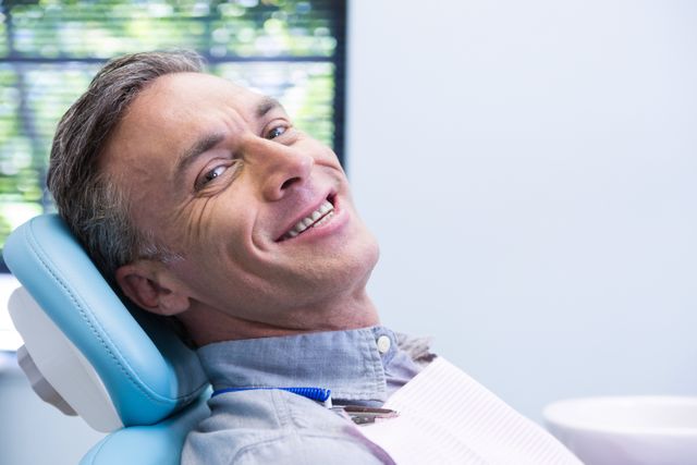 Mature man smiling while sitting in a dental chair at a clinic. Ideal for use in healthcare, dental care, and oral health promotions. Suitable for illustrating patient comfort and professional dental services.