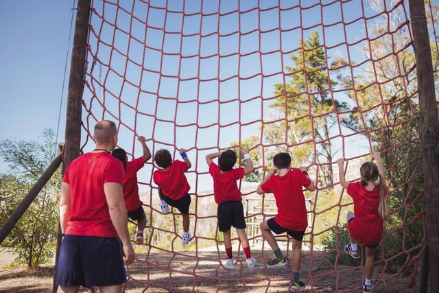 Trainer guiding group of kids climbing a net during an obstacle course training in a boot camp. Ideal for use in articles or advertisements about outdoor activities, fitness programs for children, teamwork exercises, summer camps, and physical education.