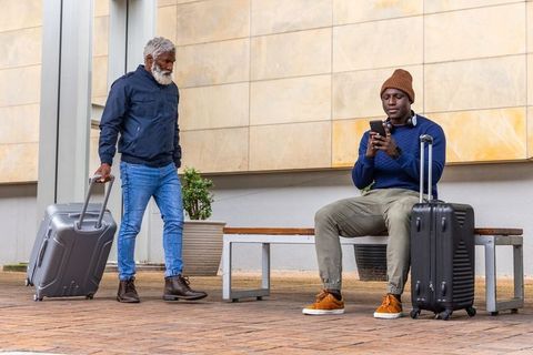 African american senior walking with suitcase while young man sitting checking phone on plaza