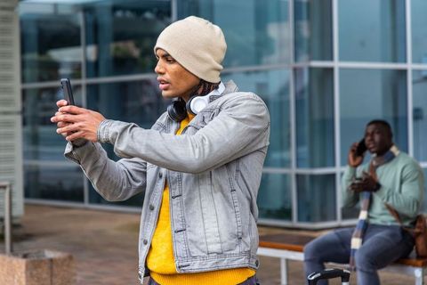 Young commuter taking phone selfie at urban transit plaza with suitcase and headphones