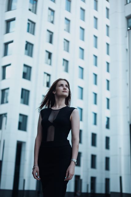Confident businesswoman dressed in elegant black outfit standing in front of a tall modern office building with reflective windows. This image can be used to depict professionalism, corporate success, or modern urban lifestyle. Suitable for business websites, promotional materials, corporate brochures, and advertising campaigns focused on professional and executive themes.