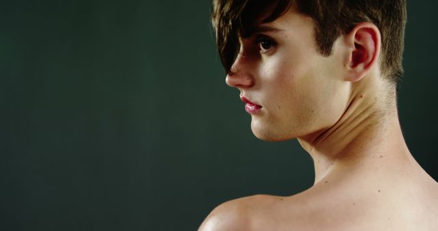 A young Caucasian individual with a short haircut is captured in a profile view against a dark background, with copy space. Their bare shoulders suggest a sense of vulnerability or the concept of natural beauty.