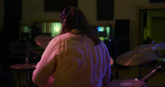 Rear view close up of a Caucasian man with long dark hair wearing headphones and playing a drum kit during a session at a recording studio. Musicians working on producing a song