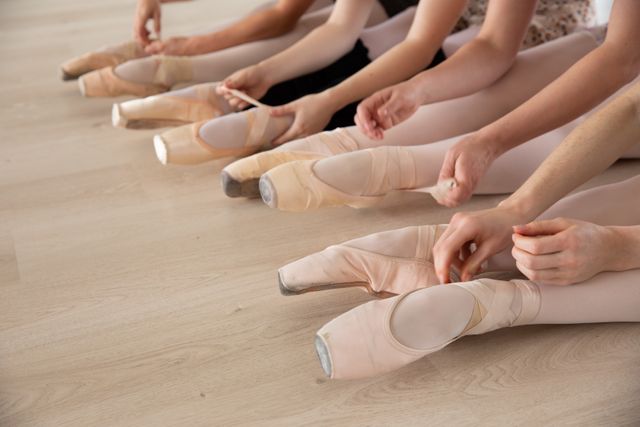roup of Caucasian female attractive ballet dancers warming up and practicing in a bright ballet studio, tying their ballet shoes sitting on the floor. Preparing for a class.