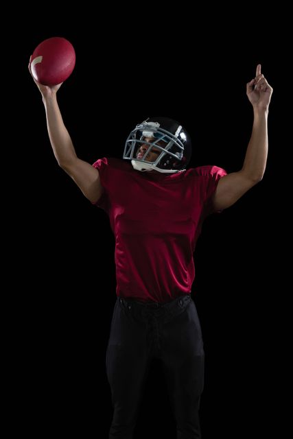 American football player raising hands holding a ball high in one hand against black background