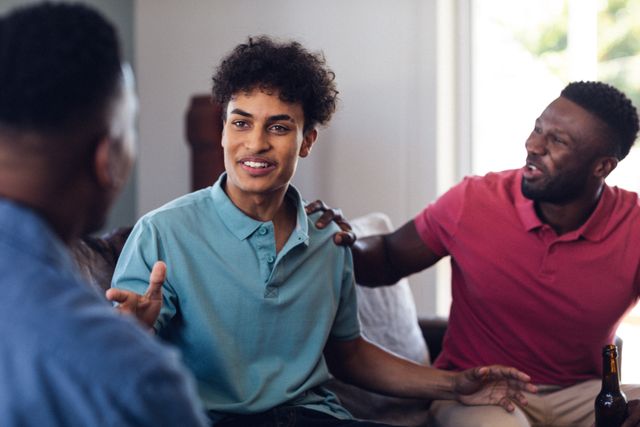 Cheerful multiracial man talking with friends while sitting on sofa in living room during weekend. Gesturing, unaltered, friendship, togetherness, social gathering, home and enjoyment concept.