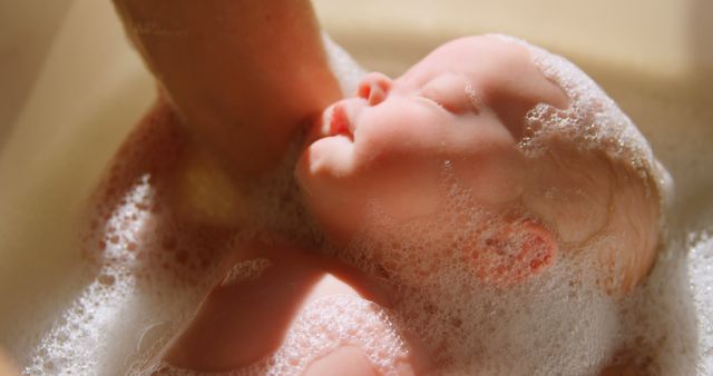 Close-up of a peaceful newborn baby being bathed with suds of bubbles. The soft lighting and focus emphasize the serenity and innocence of the child. This image is perfect for use in advertisements and articles related to parenting, baby care products, early childhood, and family life.