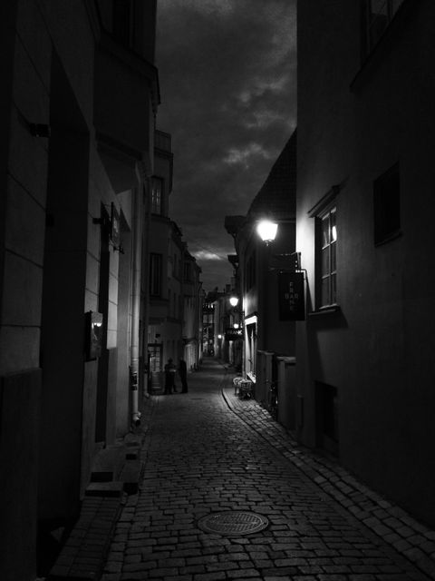 Dimly lit narrow cobblestone street at night with lamp posts illuminating historic buildings. Creates a moody, mysterious atmosphere perfect for concepts related to solitude, night walks, and historic townscapes.