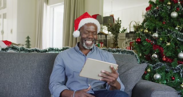 A mature man wearing a Santa hat is smiling and holding a tablet, engaging in a video call. He is seated on a gray couch in a brightly decorated living room with a Christmas tree and ornaments. This image is perfect for promoting holiday greetings, remote family celebrations, and Christmas digital communication themes.