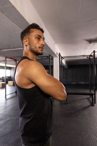 Focused athletic man standing with arms crossed in industrial gym wearing black tank top