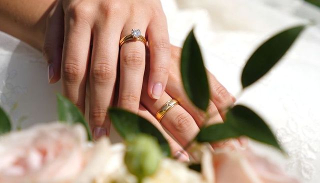 Bride's hands resting showing solitaire diamond and gold wedding band on lace dress