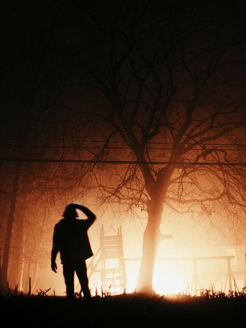 Silhouette of a person stands near a bright light under a foggy night sky, creating an eerie and dramatic scene. The light illuminates a large tree, casting long shadows across the ground. Perfect for themes related to mystery, horror, and the supernatural, as well as for use in Halloween promotions, suspenseful settings in movies, or dark-themed storytelling.