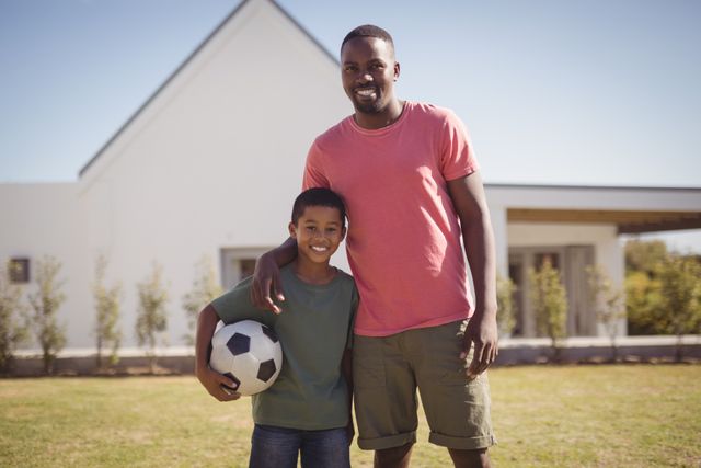 Father and son standing together in garden, smiling and holding a soccer ball. Perfect for themes related to family bonding, outdoor activities, sports, parenting, and leisure time. Ideal for use in advertisements, blogs, and articles about family life, childhood, and healthy living.