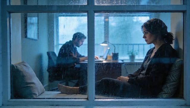 Contemplative woman staring through rainy window while partner working late on laptop