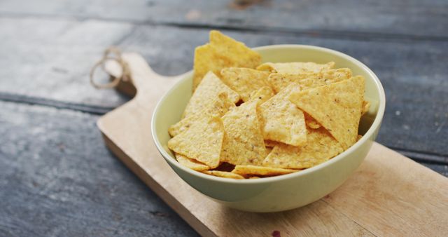 A bowl of crunchy nacho chips sits on a wooden board against a rustic blue background. This image is perfect for illustrating topics related to snack foods, parties, and casual dining. It can be used in marketing materials for restaurants, online menus, food blogs, recipe sites, and advertisements for snack products.
