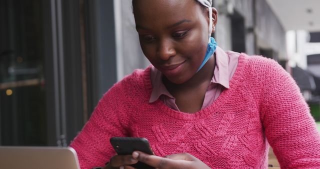 Young woman in a pink sweater smiling while using smartphone and working on laptop at outdoor cafe. Perfect for themes of technology, multitasking, student life, and everyday activities.