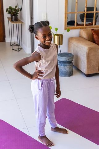 Smiling african girl standing on yoga mat in bright living room wearing lilac sweatpants