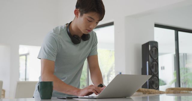 The young man is focused on typing on his laptop in a modern home. He is wearing headphones around his neck and a casual shirt, suggesting a comfortable and casual work environment. There is a coffee cup nearby, which implies he is taking a coffee break while working or studying. This image is suitable for use in articles, blogs, and marketing materials related to remote work, studying, digital nomads, technology, and lifestyle.