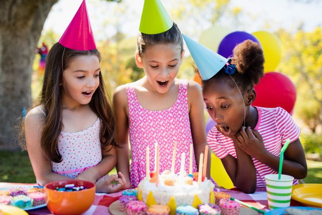 Three young girls wearing party hats are gathered around a birthday cake with lit candles, ready to blow them out. The scene is set outdoors in a park, with colorful decorations and a festive atmosphere. This image is perfect for use in advertisements, social media posts, and articles related to children's parties, celebrations, and outdoor activities.