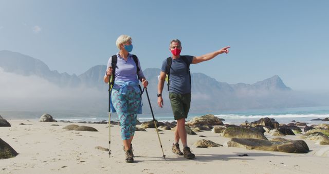 Senior hiker couple wearing face masks with backpacks and hiking poles pointing towards a direction while hiking on the beach. trekking, hiking, nature, activity, exploration, adventure concept.