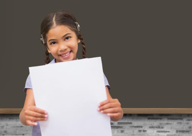 The image shows a smiling young girl with braided hair holding a blank piece of paper in a classroom setting with a blackboard in the background. Ideal for educational materials, advertisements promoting back-to-school sales, children's learning apps, and classroom decor.