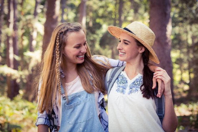 Two young women are embracing and smiling at each other while standing in a forest. They appear to be enjoying a hike or outdoor adventure, dressed in casual summer clothing. This image is perfect for promoting outdoor activities, friendship, travel, and leisure. It can be used in advertisements, social media posts, and articles related to nature, adventure, and bonding.