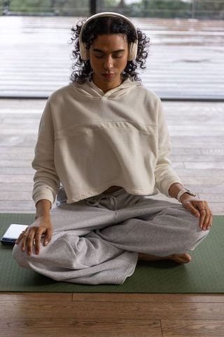 Nonbinary person meditating cross-legged on mat wearing headphones and cozy hoodie