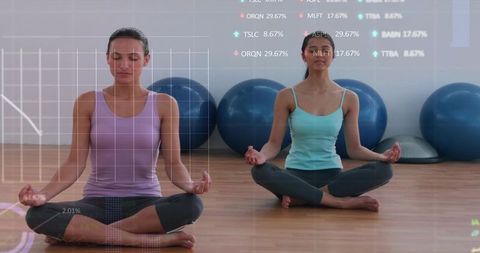 Two women meditating in yoga studio with exercise balls, bosu and data overlay for wellness