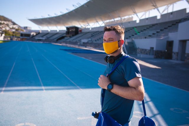 This image depicts a male athlete standing on a stadium track, wearing a face mask, headphones, and carrying a sports bag. It is ideal for use in articles or advertisements related to sports, fitness, health and safety measures during the COVID-19 pandemic, and professional athletic training.