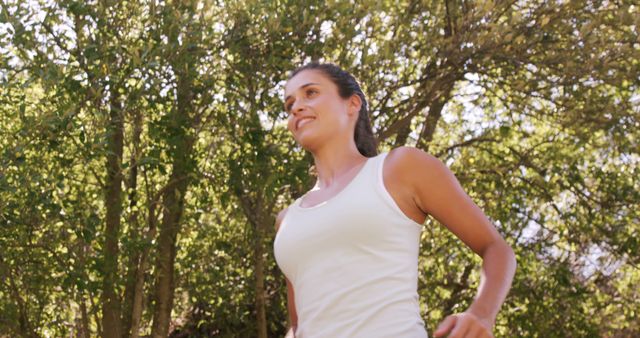Woman is jogging in outdoor forest environment with trees in the background, smiling and looking focused. Ideal for promoting topics related to health, fitness routines, outdoor activities, and active lifestyle choices.