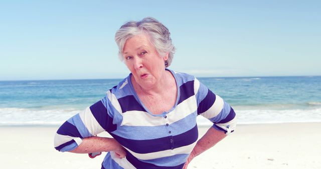 Energetic senior woman posing playfully on sunny beach with sea in background. Great for ads promoting active lifestyle, retirement, elder care, or beach vacations.