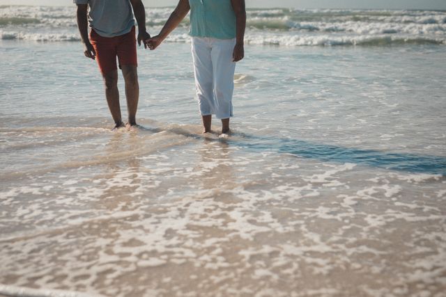 Couple holding hands while walking on the seashore with waves gently touching their feet. Ideal for themes of romance, summer vacations, beach getaways, relaxation, and togetherness. Perfect for travel brochures, romantic getaway promotions, and lifestyle blogs.