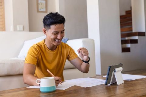 Asian young man celebrating while checking tablet and signing papers in living room