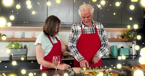 Senior couple cooking together wearing red aprons in cozy modern kitchen with wine glass