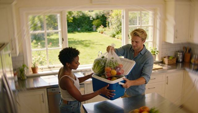 Couple unpacking groceries from cooler in bright kitchen with backyard sunlight holding fresh produc