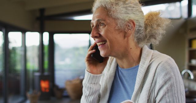 Elderly woman smiling while speaking on the phone in a cozy living room. The setting includes warm lighting and a modern interior, creating a comfortable and homely atmosphere. Perfect for ads or content related to senior lifestyle, communication, home environments, or family connections.