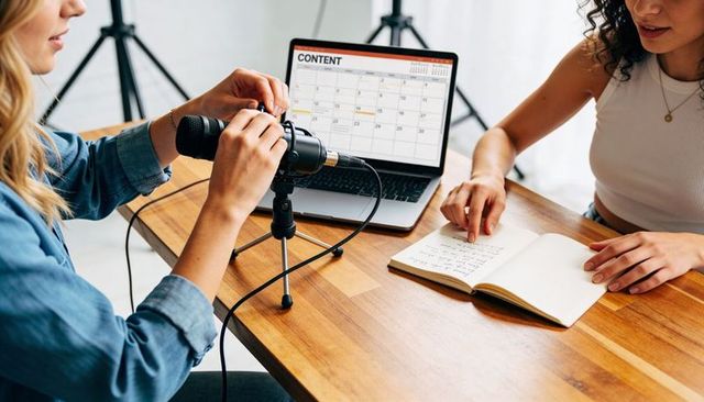Women adjusting microphone and planning content calendar on laptop for podcast recording