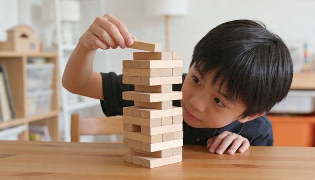 Asian boy building wooden block tower on table, practicing fine motor skills and focus