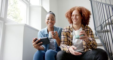 Happy Mixed-Race Couple Enjoying Coffee on Stairs with Smartphone