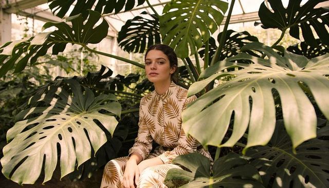 Woman wearing leaf-print outfit sitting among giant monstera leaves in glass greenhouse