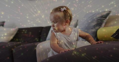 Curious toddler leaning on dark gray sofa in cozy living room with pastel bokeh lights